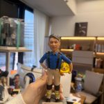 CUTEFIGUREHK 客製化榮休紀念公仔。一位灰髮男士，身穿香港消防處藍色工作服、黑色吊帶、配有護膝及反光條的卡其色長褲。他右手叉腰，左手抱著一頂印有消防徽章及「TST」字樣的黃色消防頭盔。公仔站立在仿石磚紋理的圓形灰色底座上。底座前方設有一塊專屬名牌，印有「消防總隊目 PFn11407 曾紹斌 敬業樂業33年 榮休快樂」。
