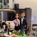客製化情侶立體真人公仔。畫面中一隻手正托著一個綠色圓形底座的雙人公仔。女方戴著黑框眼鏡，穿著深藍色無袖連身裙，雙手捧著一束粉色包裝的藍色玫瑰花；男方則穿著深藍色西裝外套、白襯衫與藍色牛仔褲，兩人面帶微笑。底座前方有一塊寫著「Happy Valentine's Day」的白色長方形銘牌。背景為公仔製作工作室，隱約可見其他公仔半成品與展示盒。