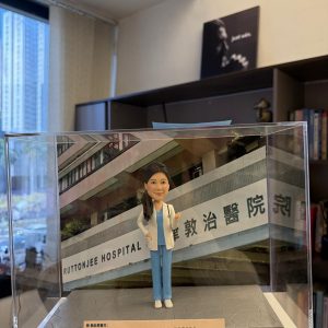 CUTEFIGUREHK 客製化女性醫生人像公仔，身穿藍色手術服和白袍並掛有聽診器。公仔放置於透明展示盒內，背景為律敦治醫院照片，底座設有刻上「仁心仁術」等真摯感謝字句的專屬牌匾，是病人送給醫生的獨一無二答謝禮物。
