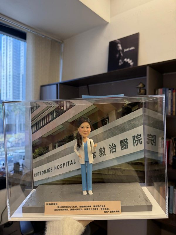CUTEFIGUREHK 客製化女性醫生人像公仔，身穿藍色手術服和白袍並掛有聽診器。公仔放置於透明展示盒內，背景為律敦治醫院照片，底座設有刻上「仁心仁術」等真摯感謝字句的專屬牌匾，是病人送給醫生的獨一無二答謝禮物。