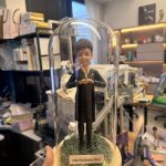CUTEFIGUREHK 客製化空服員制服公仔。短髮造型，身穿深色空姐制服配戴金色徽章，雙手比出愛心手勢。公仔放置於帶有仿真草地及LED燈串的木底座玻璃罩內，前方設有印著「ISM Christina Choi」的專屬名牌，是送給航空業人士的升職或榮休紀念禮物。