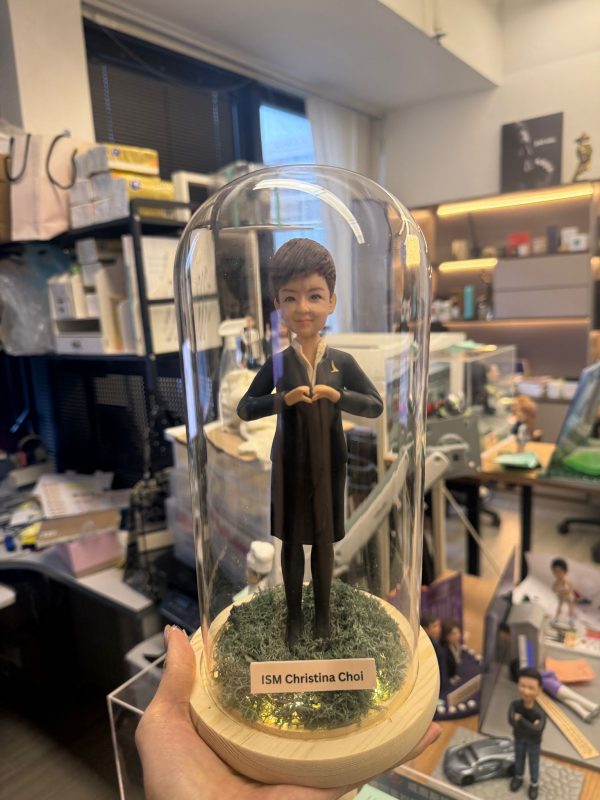 CUTEFIGUREHK 客製化空服員制服公仔。短髮造型，身穿深色空姐制服配戴金色徽章，雙手比出愛心手勢。公仔放置於帶有仿真草地及LED燈串的木底座玻璃罩內，前方設有印著「ISM Christina Choi」的專屬名牌，是送給航空業人士的升職或榮休紀念禮物。