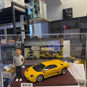 CUTEFIGUREHK 客製化人像公仔連跑車場景模型。透明展示盒內，一位穿著灰色Polo衫的男士公仔站在一輛黃色林寶堅尼跑車模型旁。背景設有「LAMBORGHINI HONG KONG」字樣的圖片背板及「告士打道」迷你路牌，底座前方印有「榮休之喜」銘牌。這是一份結合個人愛好與職業背景的專屬退休禮物。