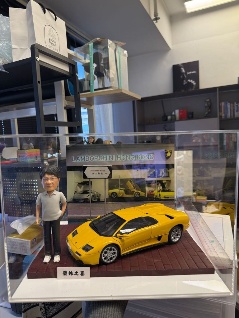 CUTEFIGUREHK 客製化人像公仔連跑車場景模型。透明展示盒內,一位穿著灰色Polo衫的男士公仔站在一輛黃色林寶堅尼跑車模型旁。背景設有「LAMBORGHINI HONG KONG」字樣的圖片背板及「告士打道」迷你路牌,底座前方印有「榮休之喜」銘牌。這是一份結合個人愛好與職業背景的專屬退休禮物。