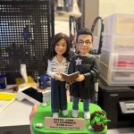 CUTEFIGUREHK 客製化雙人受洗紀念公仔。男主角穿著黑色受洗袍，女主角穿著休閒服，兩人共同捧著一本打開的聖經。公仔站在綠色草地底座上，旁邊裝飾著小花籃與白石。前方名牌印有馬可福音16:16-17金句及受洗日期「Denis So baptized 5 April 2026」，是一份充滿祝福的基督教受洗禮物。