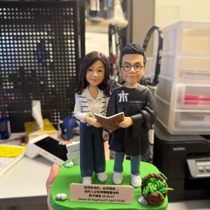 CUTEFIGUREHK 客製化雙人受洗紀念公仔。男主角穿著黑色受洗袍，女主角穿著休閒服，兩人共同捧著一本打開的聖經。公仔站在綠色草地底座上，旁邊裝飾著小花籃與白石。前方名牌印有馬可福音16:16-17金句及受洗日期「Denis So baptized 5 April 2026」，是一份充滿祝福的基督教受洗禮物。