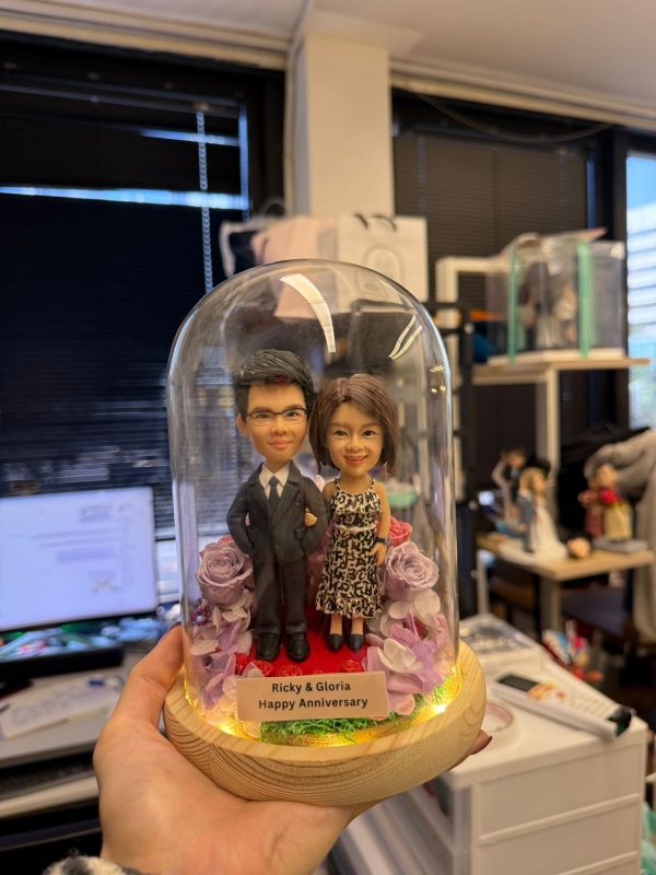 CUTEFIGUREHK 客製化情侶真人像公仔，精心佈置於帶有 LED 燈串的木座發光玻璃罩內。公仔造型為穿著西裝的男士與穿著豹紋洋裝的女士，周圍點綴著浪漫的紫色與粉色玫瑰花。底座名牌刻有「Ricky & Gloria Happy Anniversary」，是一份專為情侶或夫妻打造的浪漫週年紀念禮物。