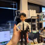 CUTEFIGUREHK 客製化男性警察人像公仔。公仔身穿香港警察經典白色制服襯衫配黑色領帶，胸前印有「POLICE 警察」字樣，左手拿著警帽，右手叉腰，展現自信英姿。公仔站立於帶有綠草與粉白小花點綴的灰色小徑圓形底座上，是一份精緻且極具紀念價值的紀律部隊禮物。