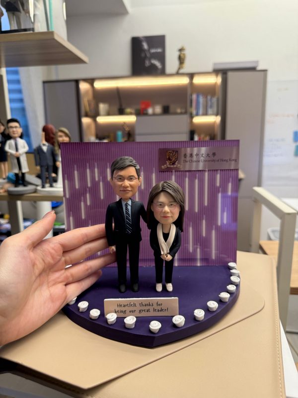 CUTEFIGUREHK 客製化雙人感恩紀念公仔。一男一女穿著專業西裝與套裝的公仔，站在以香港中文大學 (CUHK) 為背景的紫色展示台上。深紫色底座邊緣裝飾著白玫瑰，並設有寫著「Heartfelt thanks for being our great leader!」的專屬名牌，是送給上司或教授的精美告別禮物。