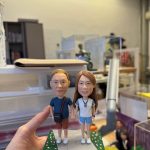 CUTEFIGUREHK 客製化情侶牽手公仔。男士穿深藍色上衣斜揹黑包，女士穿白色上衣配牛仔短褲，兩人手牽手站立於帶有綠草地與小花的灰色石板小路底座上，展現溫馨浪漫的日常散步情境，是完美的週年紀念或情人節禮物。