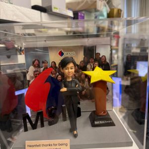 CUTEFIGUREHK 客製化女性上司人像公仔，裝在透明亞加力防塵展示盒內。公仔身穿黑色西裝，雙手交叉展現自信的領袖風範。場景內配有立體 MSIG 公司標誌及「Best Boss Award」星形獎座，底座設有印上「Heartfelt thanks for being our great leader!」的專屬木紋牌匾，背景更加入了團隊大合照，是一份極具心思的企業紀念品。