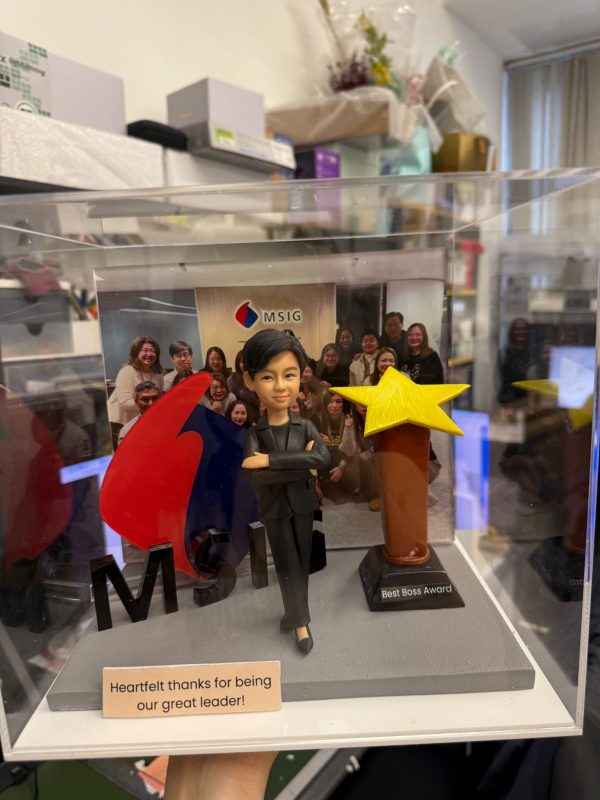 CUTEFIGUREHK 客製化女性上司人像公仔，裝在透明亞加力防塵展示盒內。公仔身穿黑色西裝，雙手交叉展現自信的領袖風範。場景內配有立體 MSIG 公司標誌及「Best Boss Award」星形獎座，底座設有印上「Heartfelt thanks for being our great leader!」的專屬木紋牌匾，背景更加入了團隊大合照，是一份極具心思的企業紀念品。
