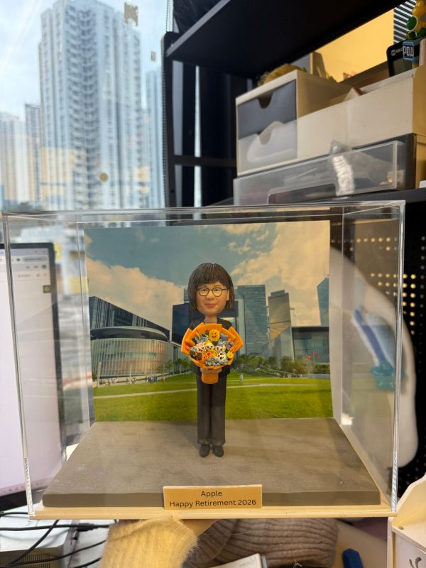CUTEFIGUREHK 客製化退休紀念真人像公仔，裝於透明壓克力展示盒內。公仔造型為穿著黑色套裝、手持繽紛花束的短髮女士。展示盒背景為香港添馬公園與政府總部大樓風景照。底座名牌刻有「Apple Happy Retirement 2026」，是一份專為慶祝榮休而訂製的專屬紀念禮物。