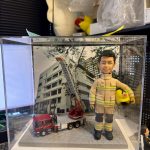 CUTEFIGUREHK 客製化消防員紀念公仔。一位身穿黃褐色消防抗火服（帶有黃色反光條紋及黑色護膝）的男士，左手抱著印有徽章的黃色消防頭盔，笑容自信。公仔放置在透明展示盒內，站在灰色石板路面設計的底座上，旁邊停泊著一輛精緻的迷你紅色消防雲梯車模型。背景板印有消防局大樓的外觀照片。這是送給消防員退休或升職的極佳紀念禮物。