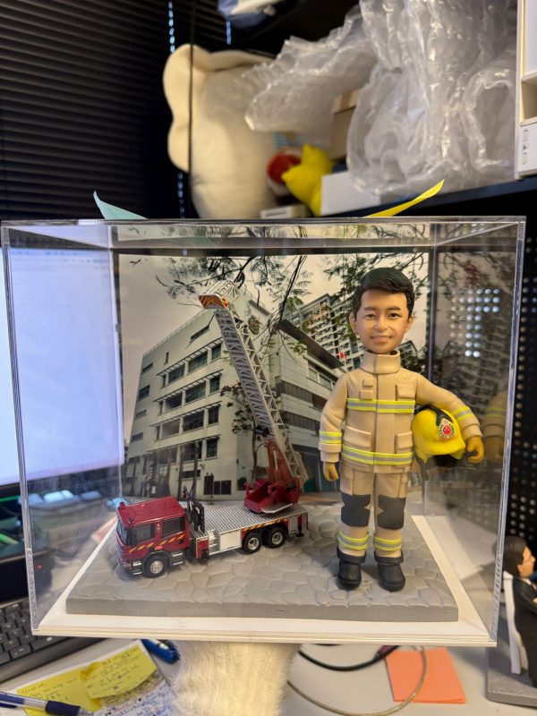 CUTEFIGUREHK 客製化消防員紀念公仔。一位身穿黃褐色消防抗火服（帶有黃色反光條紋及黑色護膝）的男士，左手抱著印有徽章的黃色消防頭盔，笑容自信。公仔放置在透明展示盒內，站在灰色石板路面設計的底座上，旁邊停泊著一輛精緻的迷你紅色消防雲梯車模型。背景板印有消防局大樓的外觀照片。這是送給消防員退休或升職的極佳紀念禮物。