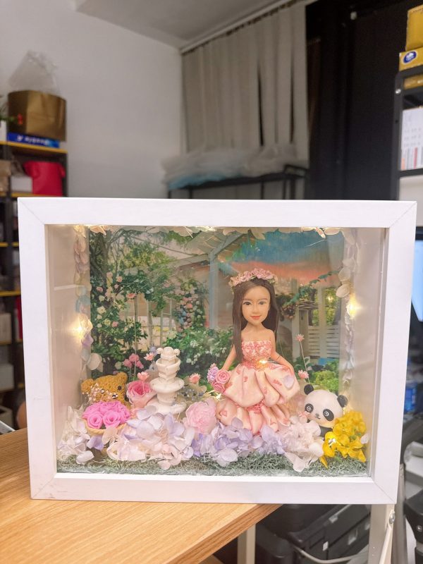 CUTEFIGUREHK 客製化立體相框公仔禮物。一個白色的發光畫框，內部佈置成夢幻的童話花園。中央是一個穿著粉紅色花瓣晚裝的長髮女孩立體公仔，頭戴花環，手持粉紅玫瑰。背景是浪漫的黃昏花園圖案，公仔腳下鋪滿了紫色、白色和粉紅色的乾花（繡球花）以及綠色苔蘚，旁邊點綴著微縮的白色小噴泉、可愛的小棕熊和熊貓模型。相框內置有溫馨的 LED 燈串，營造出唯美的氣氛。