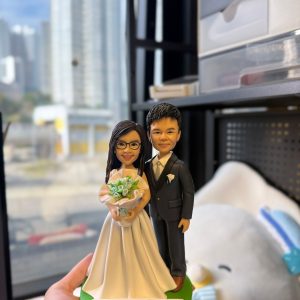 CUTEFIGUREHK 客製化夫妻結婚紀念人像公仔。女主角身穿白色婚紗、配戴眼鏡並手持淺藍色花球；男主角身穿深色西裝打領帶。綠色底座上的名牌刻有「Happy China Wedding Anniversary」的祝福語。這是一份專為慶祝結婚週年紀念度身訂造的浪漫禮物。