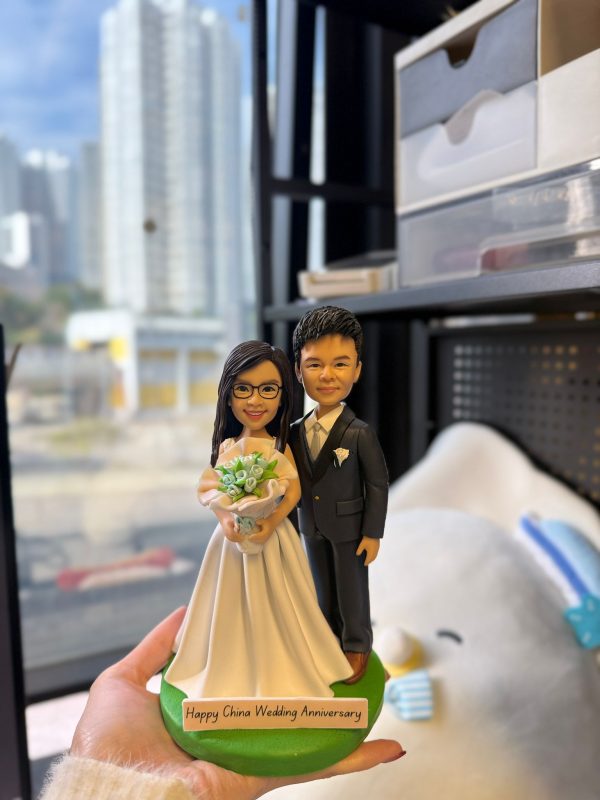 CUTEFIGUREHK 客製化夫妻結婚紀念人像公仔。女主角身穿白色婚紗、配戴眼鏡並手持淺藍色花球；男主角身穿深色西裝打領帶。綠色底座上的名牌刻有「Happy China Wedding Anniversary」的祝福語。這是一份專為慶祝結婚週年紀念度身訂造的浪漫禮物。
