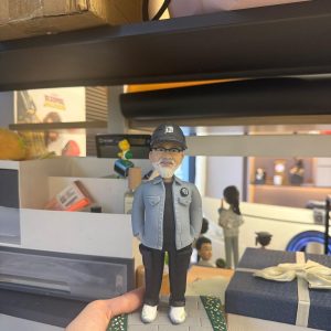 CUTEFIGUREHK 客製化男士人像公仔，生動還原了戴著黑色鴨舌帽與眼鏡、留著標誌性白鬍鬚的男士。他身穿淺藍色休閒外套、黑色上衣及工裝褲，雙手插袋，展現型格休閒風。公仔站立於帶有草地小徑的底座上，前方設有寫著「Dear David, Lululemon 10th anniversary」的專屬紀念牌。這是一份極具心思、專為慶祝員工入職十週年及長期服務而設的企業紀念禮物。