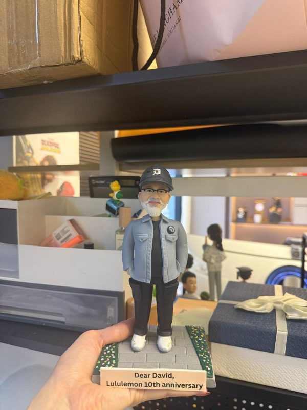 CUTEFIGUREHK 客製化男士人像公仔，生動還原了戴著黑色鴨舌帽與眼鏡、留著標誌性白鬍鬚的男士。他身穿淺藍色休閒外套、黑色上衣及工裝褲，雙手插袋，展現型格休閒風。公仔站立於帶有草地小徑的底座上，前方設有寫著「Dear David, Lululemon 10th anniversary」的專屬紀念牌。這是一份極具心思、專為慶祝員工入職十週年及長期服務而設的企業紀念禮物。