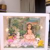 CUTEFIGUREHK 客製化立體相框公仔禮物。一個白色的發光畫框，內部佈置成夢幻的童話花園。中央是一個穿著粉紅色花瓣晚裝的長髮女孩立體公仔，頭戴花環，手持粉紅玫瑰。背景是浪漫的黃昏花園圖案，公仔腳下鋪滿了紫色、白色和粉紅色的乾花（繡球花）以及綠色苔蘚，旁邊點綴著微縮的白色小噴泉、可愛的小棕熊和熊貓模型。相框內置有溫馨的 LED 燈串，營造出唯美的氣氛。