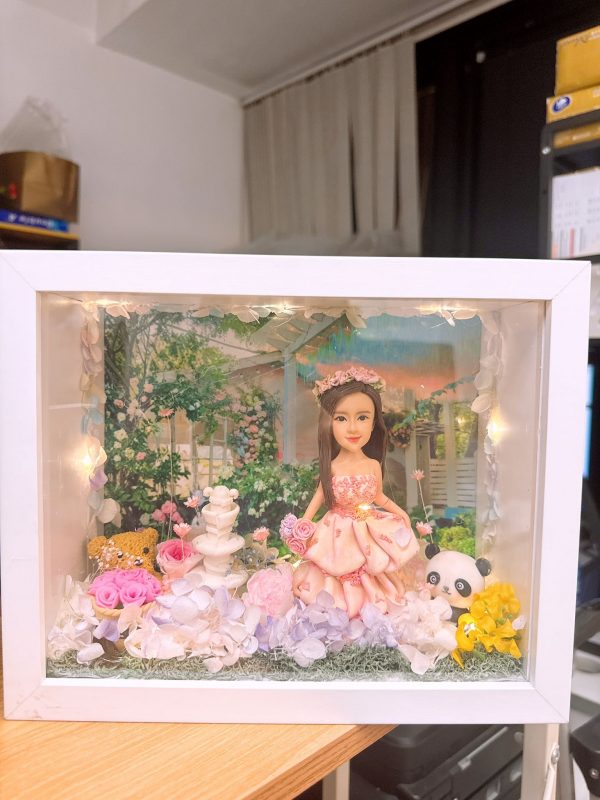 CUTEFIGUREHK 客製化立體相框公仔禮物。一個白色的發光畫框，內部佈置成夢幻的童話花園。中央是一個穿著粉紅色花瓣晚裝的長髮女孩立體公仔，頭戴花環，手持粉紅玫瑰。背景是浪漫的黃昏花園圖案，公仔腳下鋪滿了紫色、白色和粉紅色的乾花（繡球花）以及綠色苔蘚，旁邊點綴著微縮的白色小噴泉、可愛的小棕熊和熊貓模型。相框內置有溫馨的 LED 燈串，營造出唯美的氣氛。