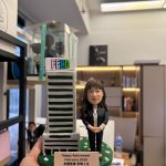 CUTEFIGUREHK 客製化榮休紀念公仔。一位穿著黑色套裝、佩戴員工證的女職員公仔，站在帶有 FEHD 標誌的微縮大樓模型旁。綠色底座點綴著花瓣，並設有寫著「Happy Retirement February 2026 身體健康 享樂人生」的專屬名牌，是專為食環署職員訂製的精美退休禮物。
