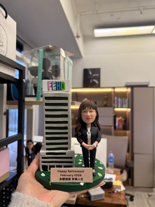 CUTEFIGUREHK 客製化榮休紀念公仔。一位穿著黑色套裝、佩戴員工證的女職員公仔，站在帶有 FEHD 標誌的微縮大樓模型旁。綠色底座點綴著花瓣，並設有寫著「Happy Retirement February 2026 身體健康 享樂人生」的專屬名牌，是專為食環署職員訂製的精美退休禮物。