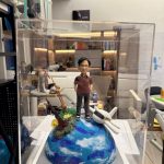 CUTEFIGUREHK 客製化環遊世界主題紀念公仔。一位戴眼鏡的男士身穿酒紅色短袖Polo衫和黑色長褲，雙手插袋。公仔站立在一個藍白相間、仿如地球表面的半球體底座上。底座上裝飾有一架白色飛機模型，以及巴黎鐵塔和東方明珠塔的微縮模型，並點綴著綠色苔蘚和黃色小花。底座前方設有一塊名牌，印有「Mr. Paul」。整個模型被放置在透明展示盒內。