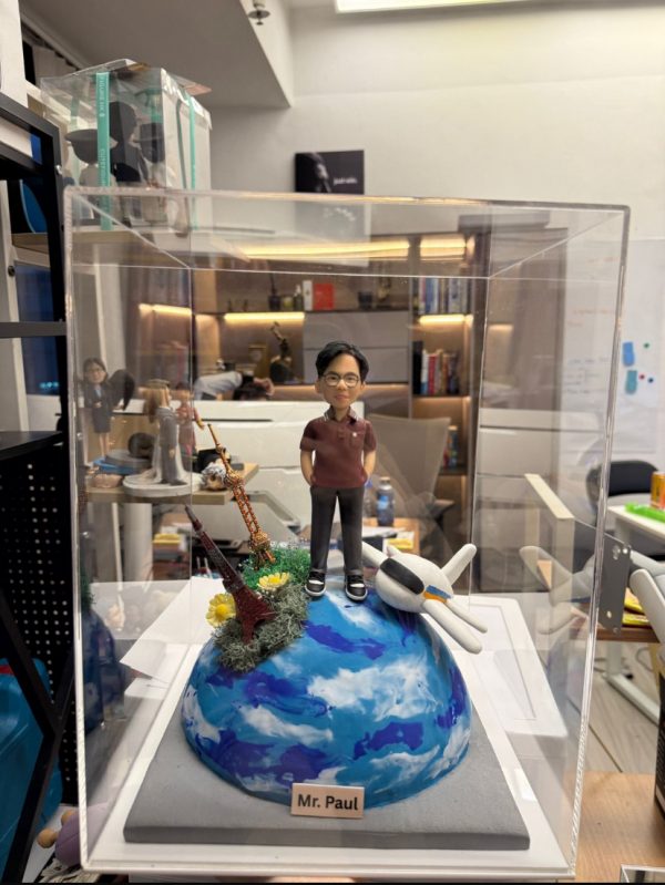 CUTEFIGUREHK 客製化環遊世界主題紀念公仔。一位戴眼鏡的男士身穿酒紅色短袖Polo衫和黑色長褲，雙手插袋。公仔站立在一個藍白相間、仿如地球表面的半球體底座上。底座上裝飾有一架白色飛機模型，以及巴黎鐵塔和東方明珠塔的微縮模型，並點綴著綠色苔蘚和黃色小花。底座前方設有一塊名牌，印有「Mr. Paul」。整個模型被放置在透明展示盒內。