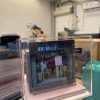 CUTEFIGUREHK 客製化店舖微縮模型。一個精緻的微型髮型屋店面模型，頂部有黑底藍字及白字的招牌寫著「Modi Hair」和「安寧路49號」。店面採用透明玻璃門設計，內部貼有價目表海報，隱約可見店內的裝潢和一張黃色高腳椅。模型放置在一個淺粉紅色的方形底座上，並罩著透明的壓克力展示盒。背景是隔著玻璃窗看到的城市高樓大廈和藍天。
