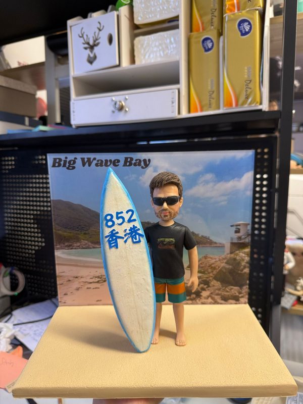 客製化單人衝浪主題真人公仔。公仔是一位留著棕色短髮與落腮鬍的男性，戴著黑色墨鏡，身穿印有波浪圖案的黑色短袖T恤與藍橘相間的衝浪褲，赤腳站立。他右手扶著一塊白底藍邊的衝浪板，板上寫有藍色的「852」與「香港」字樣。公仔站在模擬沙灘的淺黃色方形底座上，背後有一張印有「Big Wave Bay」字樣及海灘風景（包含沙灘、海浪、山丘與救生塔）的背景板。背景為室內層架與雜物。