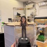 一尊裝在透明壓克力展示盒內的客製化女性公仔。公仔為長髮女性，笑容燦爛，雙手比出「YA」的手勢。她身穿深灰色短袖上衣（左胸印有「趙」字）與同色系長褲，腳穿白色厚底休閒鞋。公仔站在黑色圓形底座上，底座前方印有金色的「The Best Mum」字樣。背景為工作室環境。