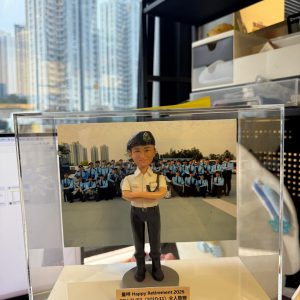 CUTEFIGUREHK 客製化男性警察人像公仔，生動還原了身穿白色短袖制服、深色長褲、黑色軍靴及配戴貝雷帽的警員造型，並擺出充滿自信的雙手交叉胸前姿勢。公仔背後配有 PTU 警察機動部隊的戶外大合照背景。底座設有專屬木紋牌匾，印有「量咩 Happy Retirement 2025 PTU Z1/E3 (2010-11) 仝人敬贈」的祝福語。這是一份極具心思的紀律部隊散水或榮休紀念禮物。