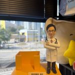 CUTEFIGUREHK 客製化醫生禮物公仔。一個穿著白袍、戴著黑框眼鏡和聽診器的男醫生立體公仔，雙手交叉抱胸，展現專業自信的笑容。公仔站在方形黃色底座上，旁邊配有一個實用的黃色名片座（或筆筒）設計。底座前方鑲有一塊客製化銘牌，上面印有「黃劍明醫生 惠存 仁心仁術 方松洲家人 敬贈」的致謝字句。背景為窗外的城市街景。