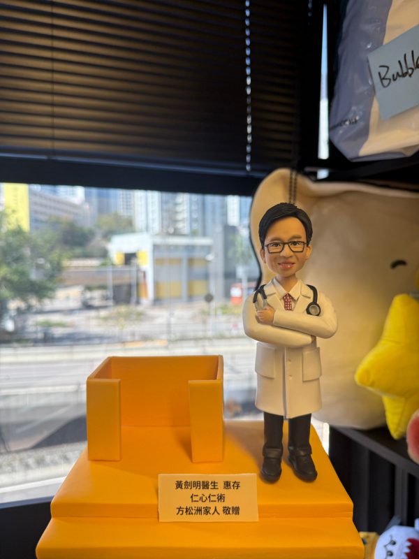 CUTEFIGUREHK 客製化醫生禮物公仔。一個穿著白袍、戴著黑框眼鏡和聽診器的男醫生立體公仔，雙手交叉抱胸，展現專業自信的笑容。公仔站在方形黃色底座上，旁邊配有一個實用的黃色名片座（或筆筒）設計。底座前方鑲有一塊客製化銘牌，上面印有「黃劍明醫生 惠存 仁心仁術 方松洲家人 敬贈」的致謝字句。背景為窗外的城市街景。