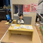 CUTEFIGUREHK 客製化退休禮物公仔。一個精緻的辦公室場景立體模型，主角是一位短髮女士，身穿黑白格紋外套，雙手交叉自信地坐在辦公桌前。黃色的辦公桌上放滿了文件和一部白色電話。旁邊的側桌有一部顯示 Windows 11 畫面的電腦屏幕及鍵盤滑鼠。背景牆上有窗戶透出城市高樓景色，窗台放有打印機，牆上貼有「2025 香港義工獎」及「國際義工日」的縮微海報。模型前方底座有一塊紅色名牌，用白字寫著「Goodbye documents, Hello Husband」及「Likie Lee CEO」。