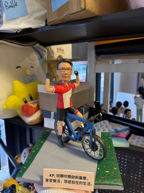 客製化單人自行車真人公仔。公仔是一位戴著黑框眼鏡的男性，身穿紅底白塊的短袖上衣、黑色單車褲與黑色半指手套。他跨騎在一輛藍色公路自行車上，雙手握拳高舉，擺出充滿活力與自信的慶祝姿勢。公仔固定在一個模擬灰色柏油路與兩側綠色草地（點綴著彩色小花）的方形底座上。底座前方有一塊白色字牌，上面印有中文字：「KP, 祝願你開啟新篇章，享受慢活，悠遊自在的生活。」。背景為擺放雜物的層架。