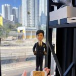 CUTEFIGUREHK 客製化飛機師禮物公仔。一隻手托著一個精緻的立體真人公仔，背景是城市高樓與公路的窗外日景。公仔主角是一位身穿深藍色飛機師制服的男士，制服袖口有金色條紋，內搭白襯衫與圖案領帶，雙手插袋，神態自信從容。公仔站在白色的圓形底座上，底座前方有一塊黃色名牌，上面用黑字寫著英文勵志金句「Never quit. Never give up. Fly it to the end.」。