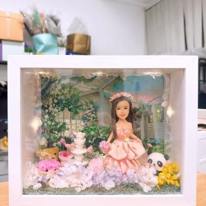 CUTEFIGUREHK 客製化立體相框公仔禮物。一個白色的發光畫框，內部佈置成夢幻的童話花園。中央是一個穿著粉紅色花瓣晚裝的長髮女孩立體公仔，頭戴花環，手持粉紅玫瑰。背景是浪漫的黃昏花園圖案，公仔腳下鋪滿了紫色、白色和粉紅色的乾花（繡球花）以及綠色苔蘚，旁邊點綴著微縮的白色小噴泉、可愛的小棕熊和熊貓模型。相框內置有溫馨的 LED 燈串，營造出唯美的氣氛。