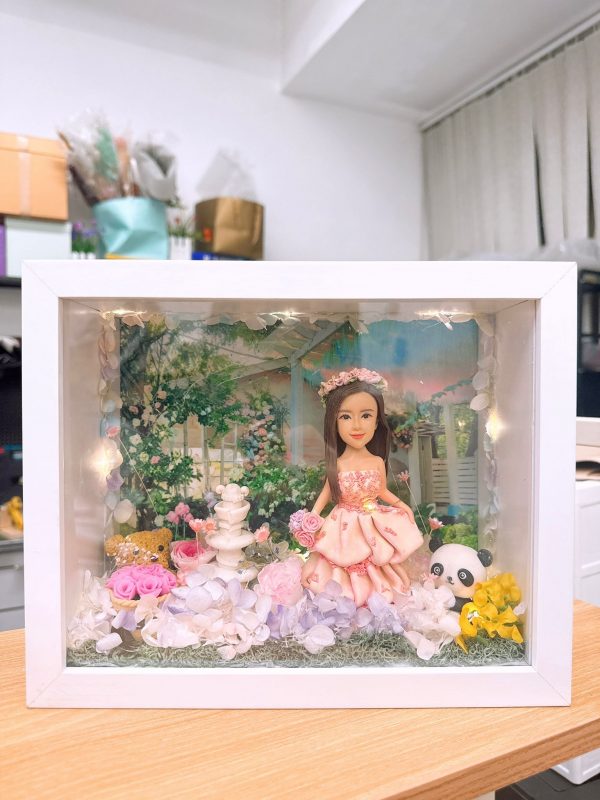 CUTEFIGUREHK 客製化立體相框公仔禮物。一個白色的發光畫框，內部佈置成夢幻的童話花園。中央是一個穿著粉紅色花瓣晚裝的長髮女孩立體公仔，頭戴花環，手持粉紅玫瑰。背景是浪漫的黃昏花園圖案，公仔腳下鋪滿了紫色、白色和粉紅色的乾花（繡球花）以及綠色苔蘚，旁邊點綴著微縮的白色小噴泉、可愛的小棕熊和熊貓模型。相框內置有溫馨的 LED 燈串，營造出唯美的氣氛。