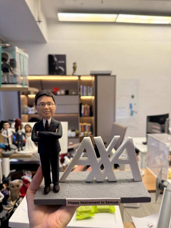 CUTEFIGUREHK 客製化男士退休公仔。公仔身穿筆挺黑色西裝、戴眼鏡並雙手交叉胸前，展現專業自信。旁邊配有大型立體 AXA 企業標誌，灰色仿石紋底座前方印有「Happy Retirement to Samuel」專屬祝賀語名牌，是完美的企業榮休紀念禮物。