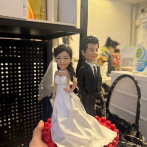 CUTEFIGUREHK 客製化新郎新娘結婚公仔。新娘身穿優雅白色露肩婚紗配戴頭紗，新郎身穿筆挺黑色西裝打領帶。雙人公仔站立於佈滿紅玫瑰裝飾的精緻紅色心形底座上，適合用作結婚蛋糕裝飾 (Cake Topper) 或專屬結婚禮物。