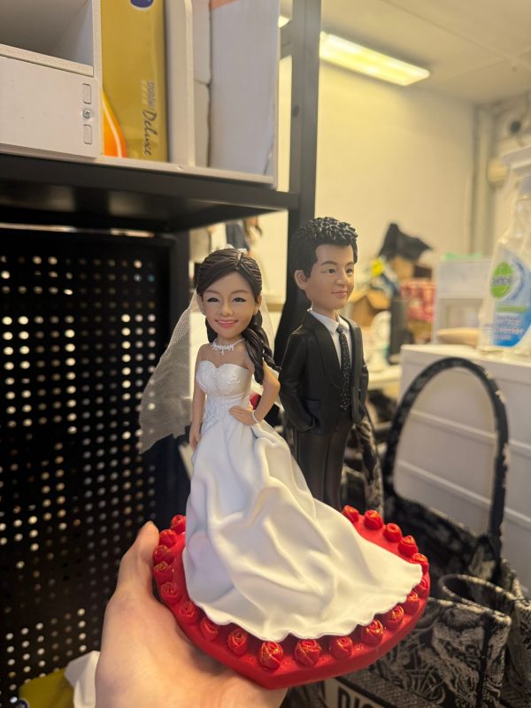 CUTEFIGUREHK 客製化新郎新娘結婚公仔。新娘身穿優雅白色露肩婚紗配戴頭紗，新郎身穿筆挺黑色西裝打領帶。雙人公仔站立於佈滿紅玫瑰裝飾的精緻紅色心形底座上，適合用作結婚蛋糕裝飾 (Cake Topper) 或專屬結婚禮物。