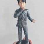 客製化單人立體真人公仔。一隻手托著一個黑色圓形底座的男性公仔。公仔有著黑色的旁分短髮，五官清秀。他穿著一套引人注目的銀灰色菱格紋西裝套裝，搭配黑白相間的休閒帆布鞋。公仔右手輕觸下巴，左手向外微攤，擺出自信且充滿魅力的姿勢。背景為純白牆面。