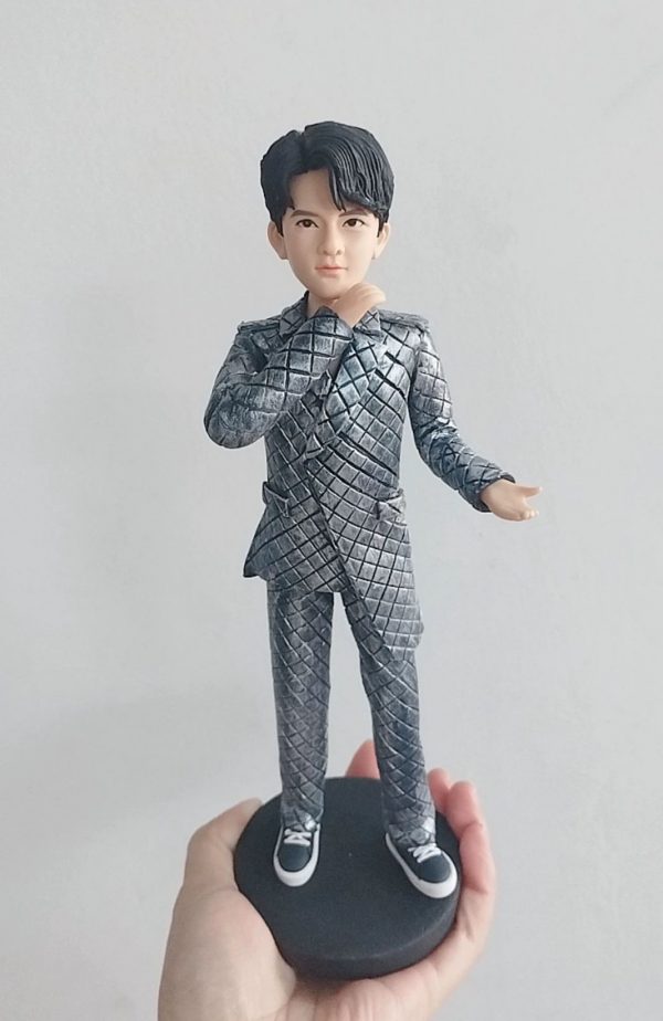 客製化單人立體真人公仔。一隻手托著一個黑色圓形底座的男性公仔。公仔有著黑色的旁分短髮，五官清秀。他穿著一套引人注目的銀灰色菱格紋西裝套裝，搭配黑白相間的休閒帆布鞋。公仔右手輕觸下巴，左手向外微攤，擺出自信且充滿魅力的姿勢。背景為純白牆面。