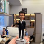 CUTEFIGUREHK 客製化男士公仔。人偶穿著深色西裝外套配白襯衫與白球鞋，展現商務休閒風格。右手舉起食指，面帶自信微笑。灰色圓形底座上帶有「Louis Antoine」專屬名牌及小金星圖案，是完美的升職或職場紀念禮物。