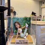 CUTEFIGUREHK 客製化瑜伽主題紀念公仔。一位留著棕色中長髮的女士，身穿白色運動內衣、淺色瑜伽褲及帶有紅白藍條紋的白襪，正在木質底座上優雅地展現單腿伸展的瑜伽姿勢。底座前方設有一塊專屬名牌，印有名字「Mei Yan」及瑜伽哲理金句「瑜伽止息心念波動」。公仔背後配有一幅綠意盎然的自然植物背景圖，整體安放在一個透明的亞加力膠展示盒內。
