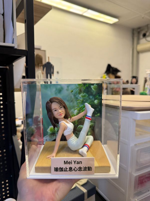 CUTEFIGUREHK 客製化瑜伽主題紀念公仔。一位留著棕色中長髮的女士，身穿白色運動內衣、淺色瑜伽褲及帶有紅白藍條紋的白襪，正在木質底座上優雅地展現單腿伸展的瑜伽姿勢。底座前方設有一塊專屬名牌，印有名字「Mei Yan」及瑜伽哲理金句「瑜伽止息心念波動」。公仔背後配有一幅綠意盎然的自然植物背景圖，整體安放在一個透明的亞加力膠展示盒內。