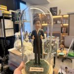 CUTEFIGUREHK 客製化空服員制服公仔。盤髮造型，身穿深色空姐制服配戴金色徽章，做出讚好手勢。公仔放置於帶有仿真草地的木底座玻璃罩內，前方設有印著「ISM Kimmy Chan」的專屬名牌，是送給航空業人士的升職或榮休紀念禮物。